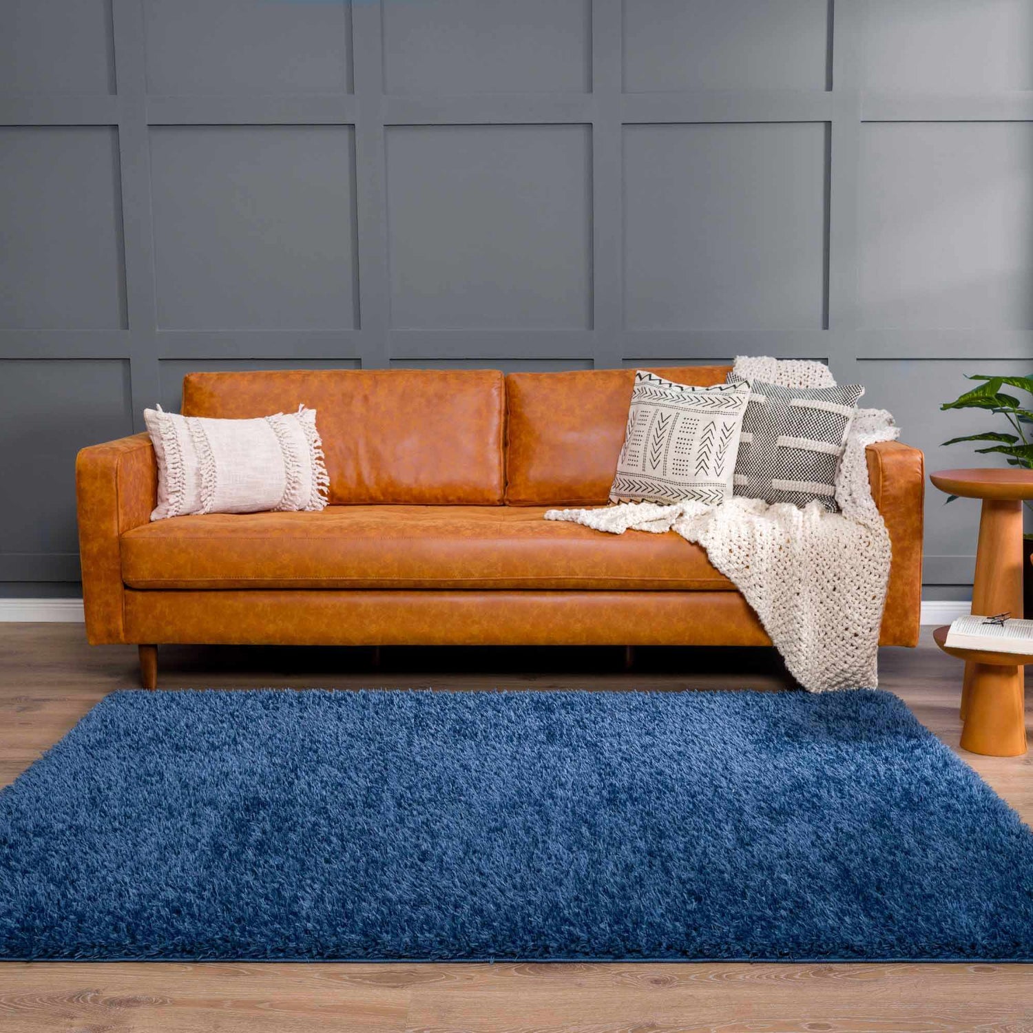 Living room scene with a Faina Solid Dark Blue Shag rug, tan leather sofa, patterned pillows, and wood accents. Roomscene-Rectangle