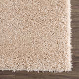 Close-up of the plush texture of the Faina Solid Beige Shag Washable Area Rug on dark wood flooring. Swatch