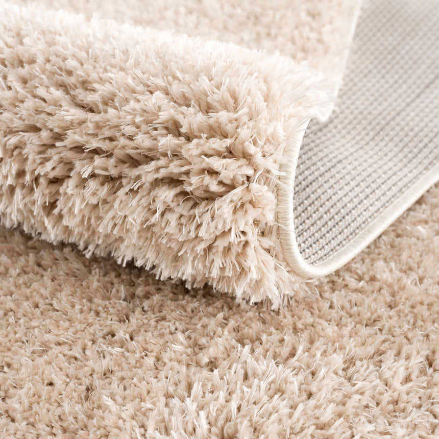 Close-up of the plush Faina Solid Beige Shag Rug, showing its soft texture and woven backing. Fold