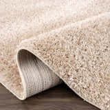Close-up of the plush texture and backing of a Faina Solid Beige Shag Washable Area Rug on a dark wood floor. Fold
