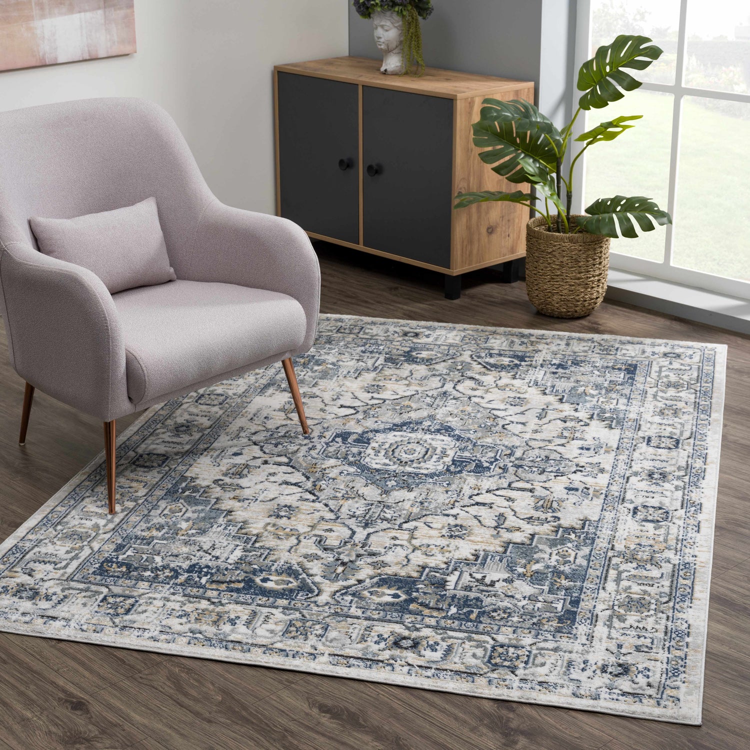 Living room scene with a blue, cream, and gold Murrindindi Area Rug, light gray chair, and wood cabinet with plant. Roomscene-Rectangle