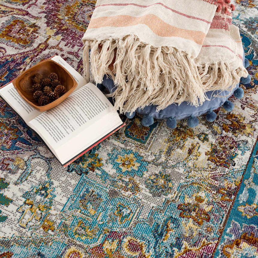 Colorful Mardin area rug detail with book, pinecones, and throw blanket. Bohemian style rug pattern in blue, pink, and mustard. Styleshot