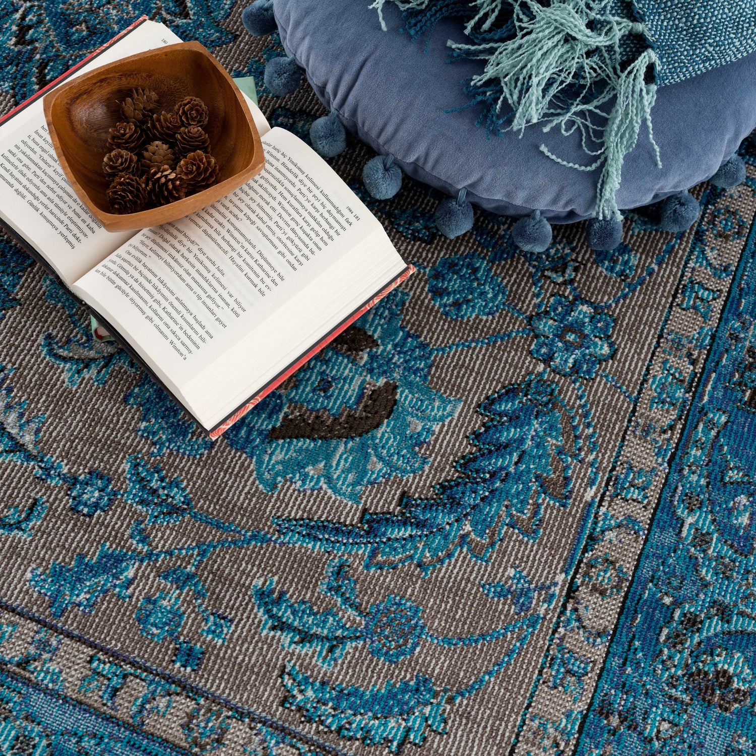 Close-up of the beige and turquoise Mardina Turquoise Area Rug with a book, pinecones, and a blue pillow. Styleshot