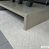 Close-up of the cream Maribyrnong 3D Outdoor Area Rug featuring a raised, geometric pattern under a gray concrete coffee table. Roomscene-Rectangle