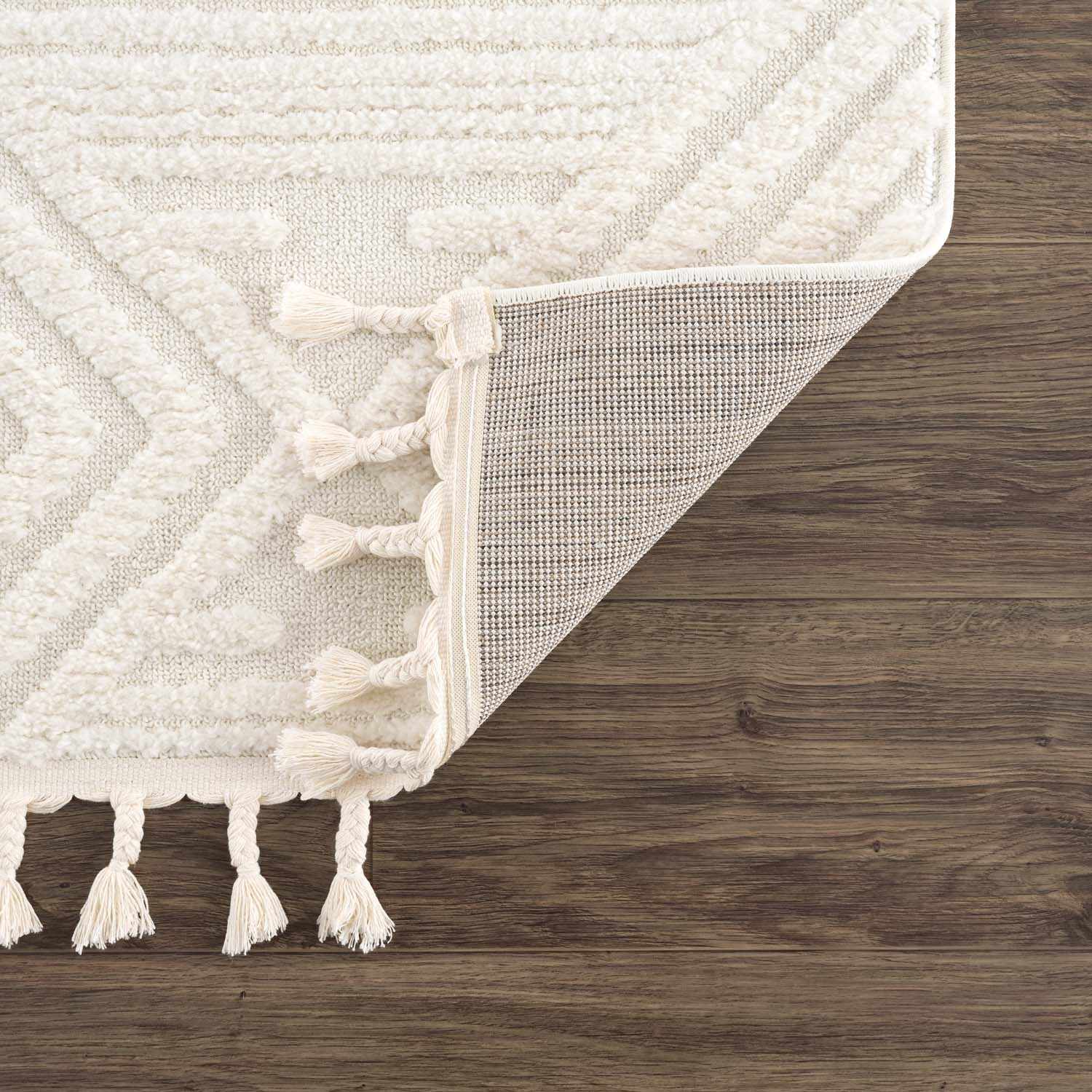 Close-up of the Manyas 3D Bone Area Rug showing geometric pattern, tassels, and texture on dark wood floor. Fold