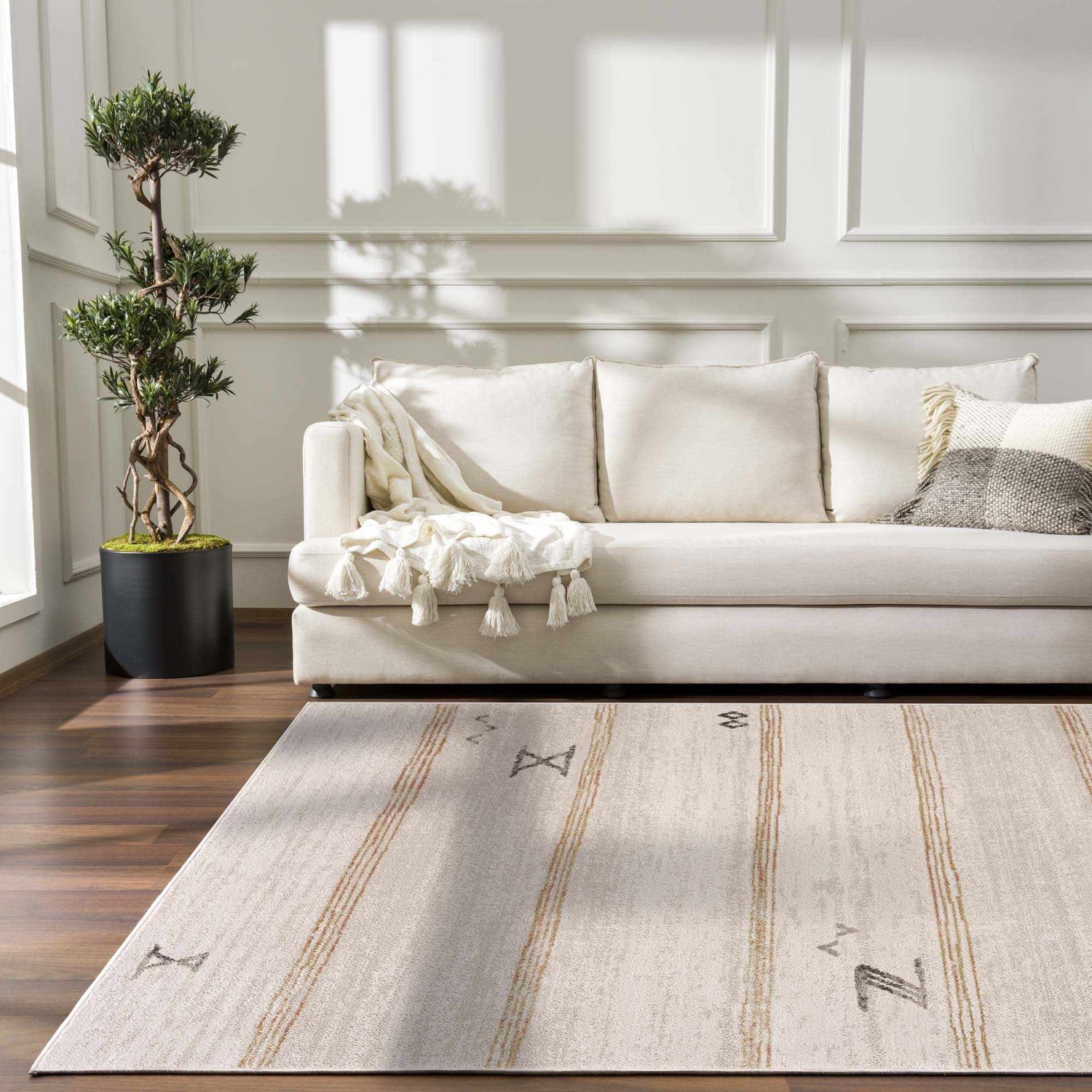 Living room scene with a Deja Cream & Gold Area Rug, cream sofa with throw, dark wood floor, and potted tree. Roomscene-Rectangle