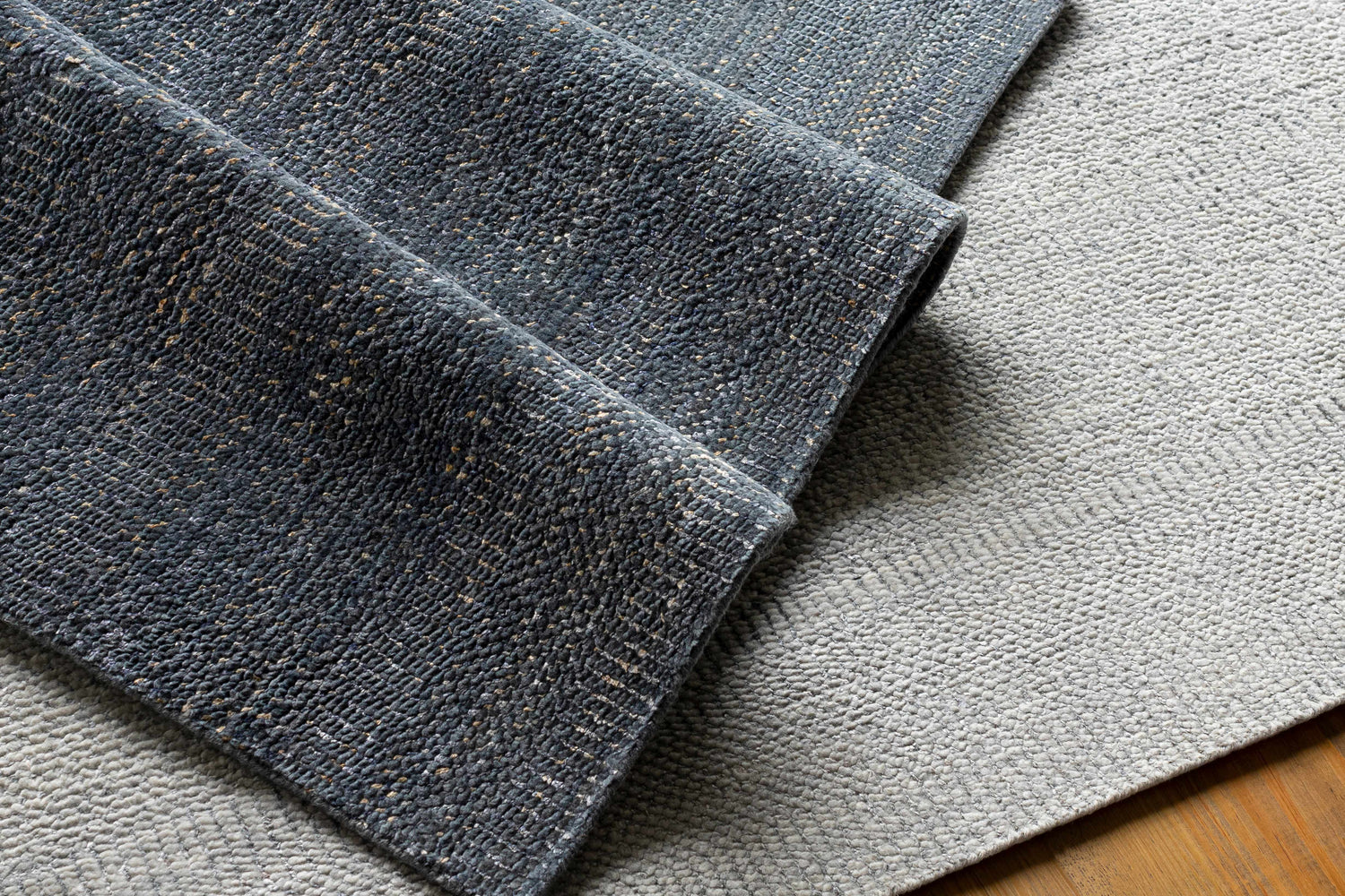 Close-up of the hand-knotted Hoole Area Rug. Dark blue wool rug with tan accents, folded over an off-white textured rug. Styleshot