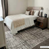 Bedroom scene with a dark gray and taupe Taysan Area Rug under a bed with neutral bedding and a wood nightstand. Roomscene-Rectangle