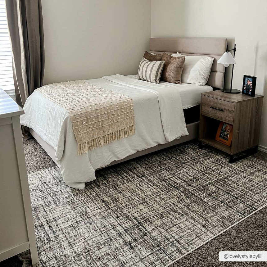 Bedroom scene with a dark gray and taupe Taysan Area Rug under a bed with neutral bedding and a wood nightstand. Roomscene-Rectangle