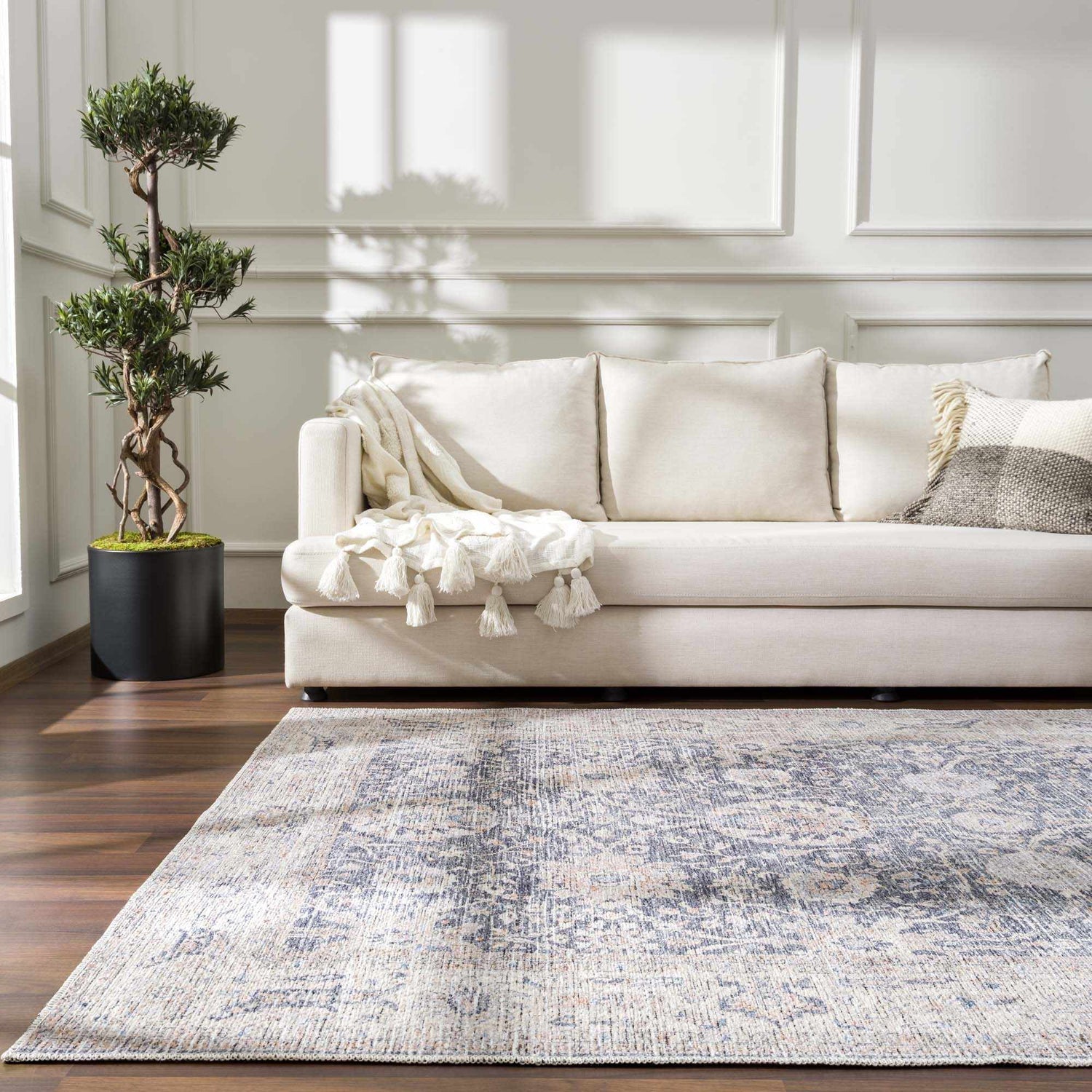 Living room scene with a silver and gray Kayin Washable Rug, cream sofa, wood floors, and a potted tree. Roomscene-Rectangle