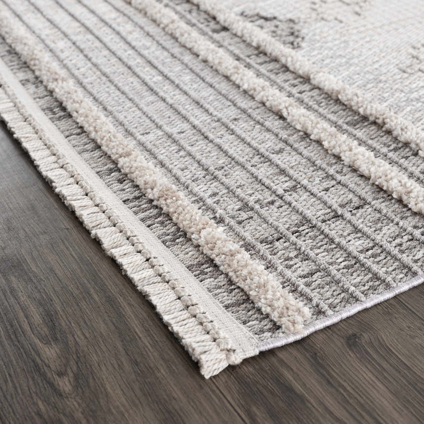 Close up of the Amaga High-Low Rustic Rug. Charcoal, cream, and gray geometric pattern with high-low texture and fringe detail. Corner