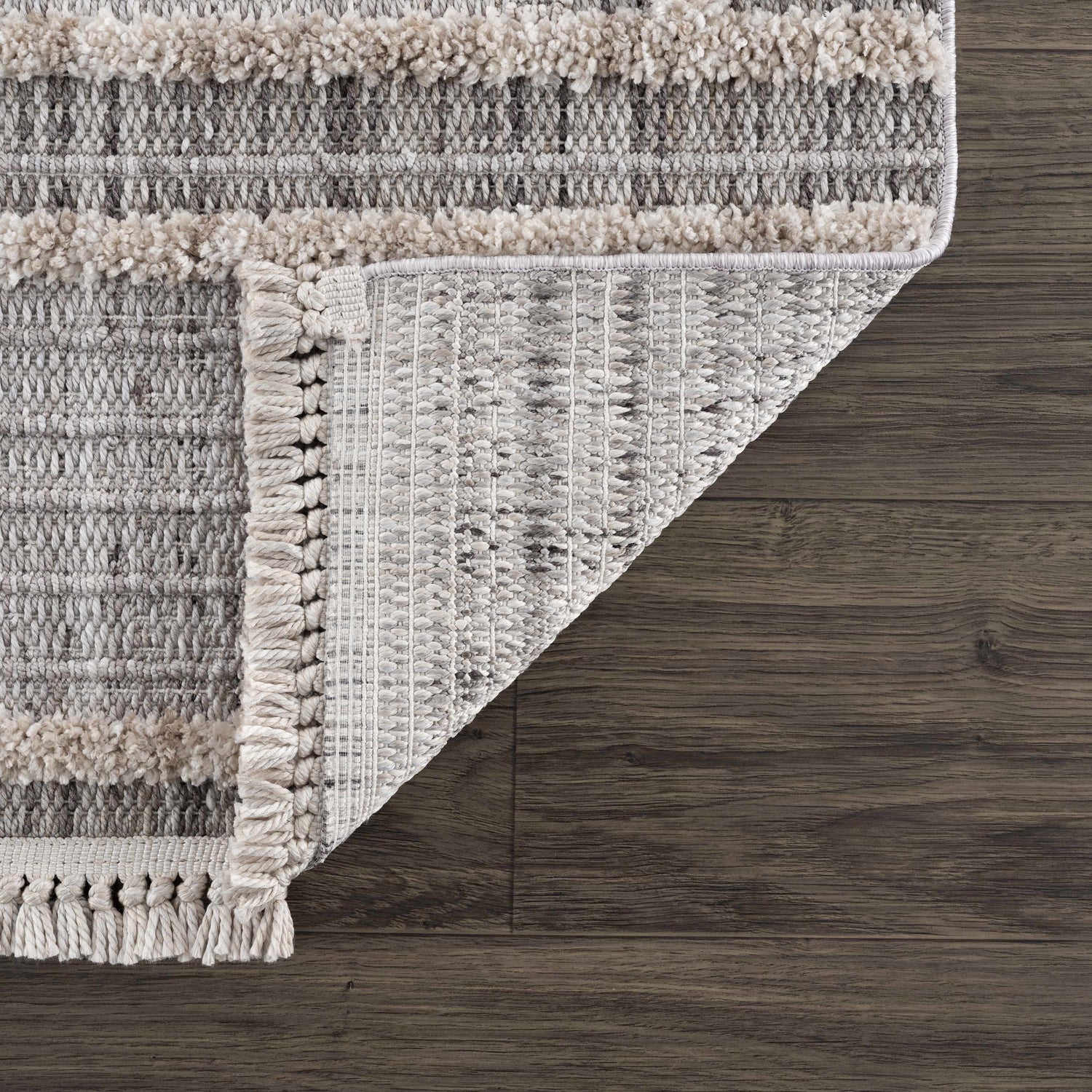 Close-up of the Amaga High-Low Rustic Rug showing texture, geometric pattern, fringe, and color palette of charcoal, cream, and gray. Fold
