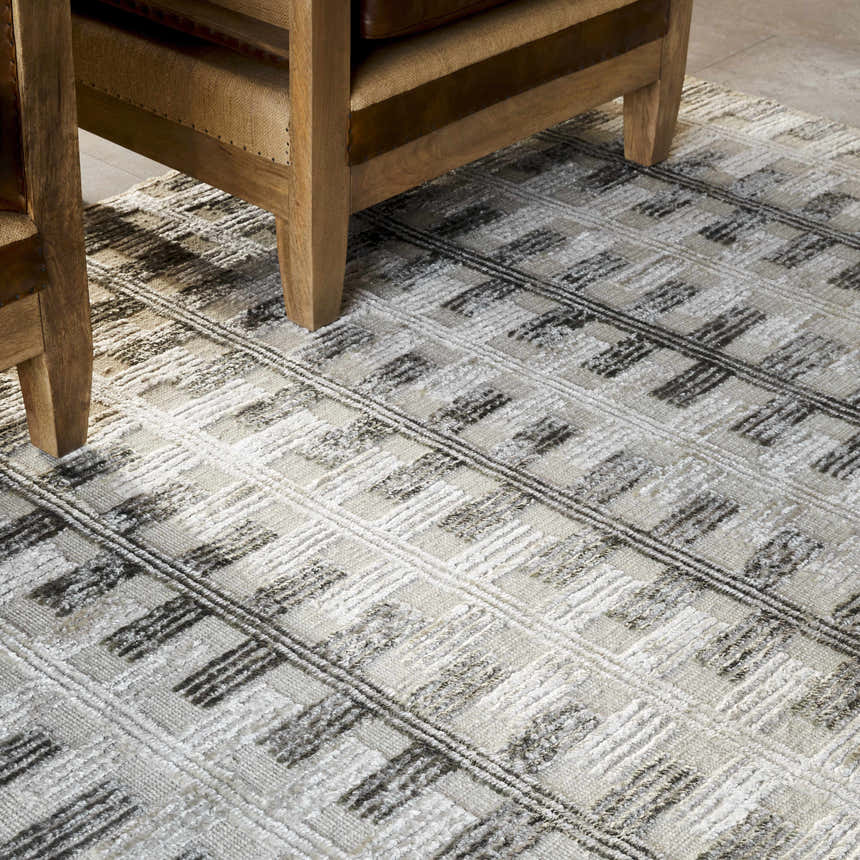 Close-up of the Aldis Grey Plush Textured Washable Rug featuring a geometric pattern and neutral colors under a wood chair. Styleshot