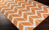Close up of Amalia Area Rug showing orange and cream geometric chevron pattern on dark wood floor. Corner