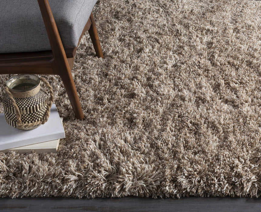 Close-up of the beige Ascot Area Rug, a hand-woven shag rug. Features a gray chair and decorative vase on books. Styleshot