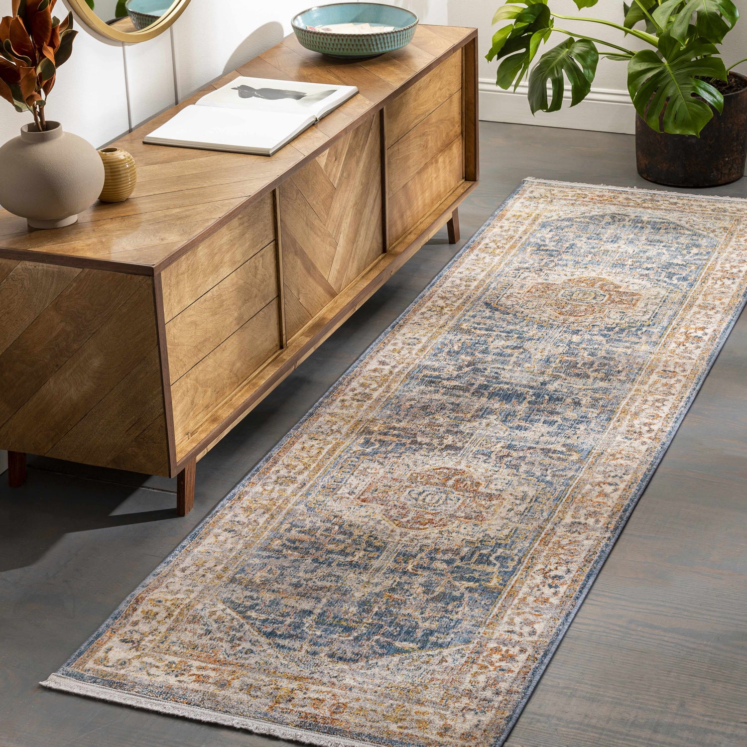 Hallway scene with a Solana Distressed Medallion Area Carpet in blue, orange, and cream colors. Wood cabinet and monstera plant. Roomscene-Runner