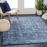 Living room scene with the Michie Dark Blue Area Rug, woven chair, blue pillow, and white pouf. Roomscene-Rectangle
