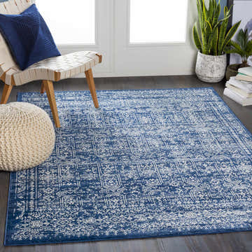 Living room scene with the Michie Dark Blue Area Rug, woven chair, blue pillow, and white pouf. Roomscene-Rectangle
