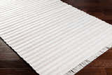 Close-up of the light gray Benek Area Rug showing its geometric, hand-woven design and fringe detail on dark wood floors. Corner