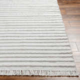 Close-up of light gray Benek Area Rug showing hand woven texture, geometric pattern, and fringe detail on wood floor. Front