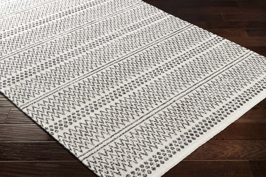 Close-up of Bewbush Washable Area Rug. Cream cotton rug with black bohemian pattern on dark wood floor. Corner