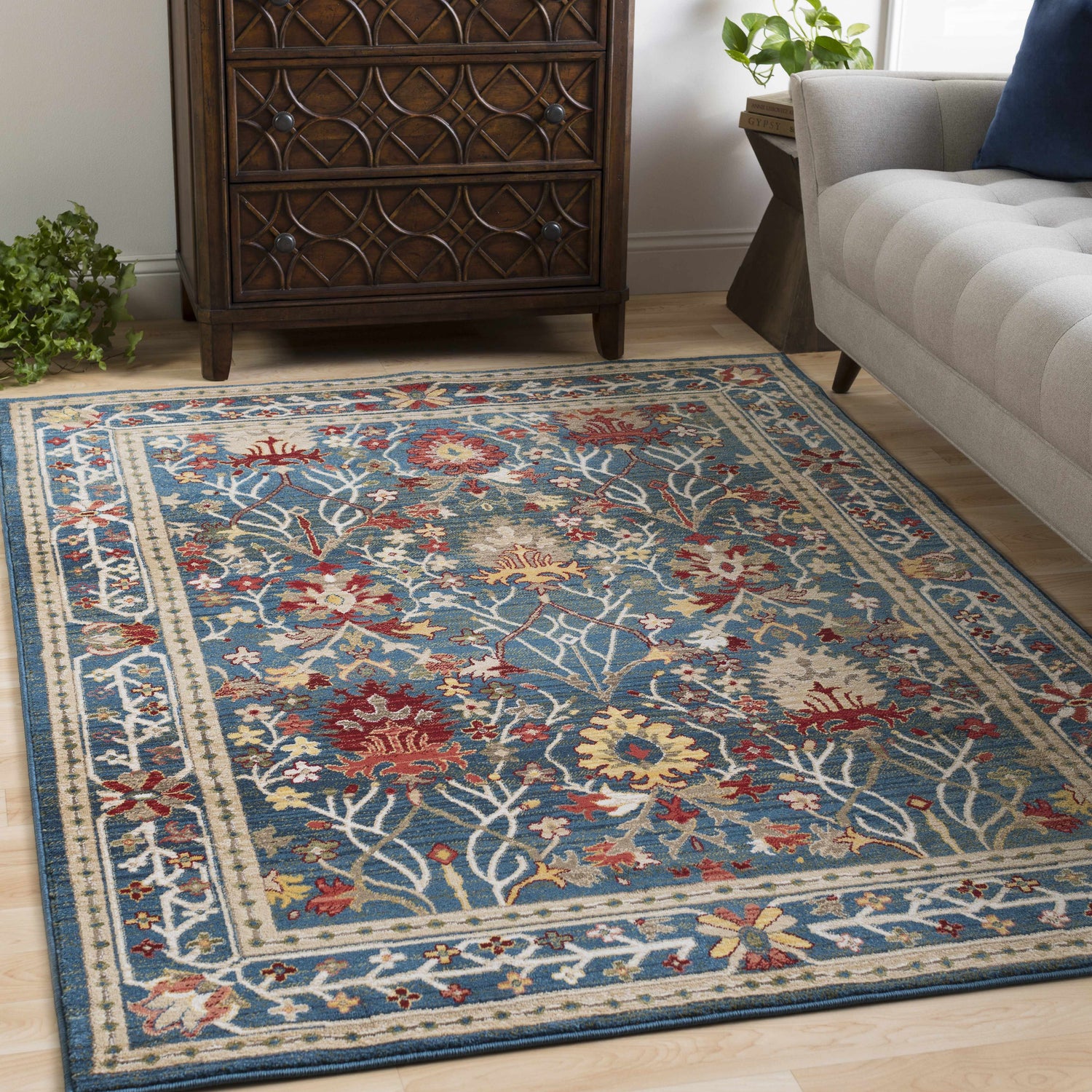 Living room scene with a Neola Navy Blue Damask Area Rug, wood dresser, and gray sofa. Floral pattern in red, yellow, and tan. Roomscene-Rectangle