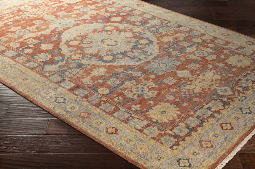 Close-up of the hand-knotted Brookeland Area Rug, a wool rug in beige, brick red, and blue, on dark wood flooring. Corner