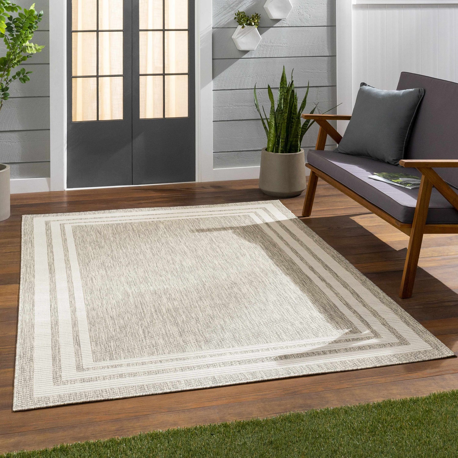 Outdoor patio scene with a beige Burradoo Bordered Area Rug, gray loveseat, wood floors, and potted plants. Roomscene-Rectangle