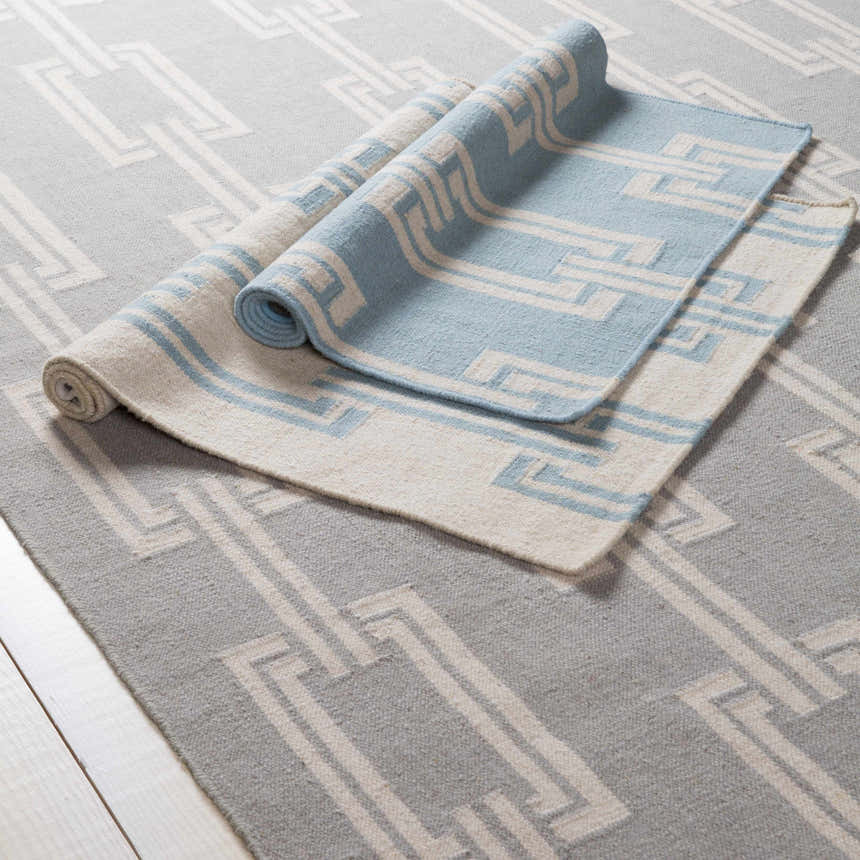 Close-up of Brundidge Area Carpet. Hand-woven wool rug in gray and ivory with contemporary geometric pattern. Runner rug partially rolled. Styleshot