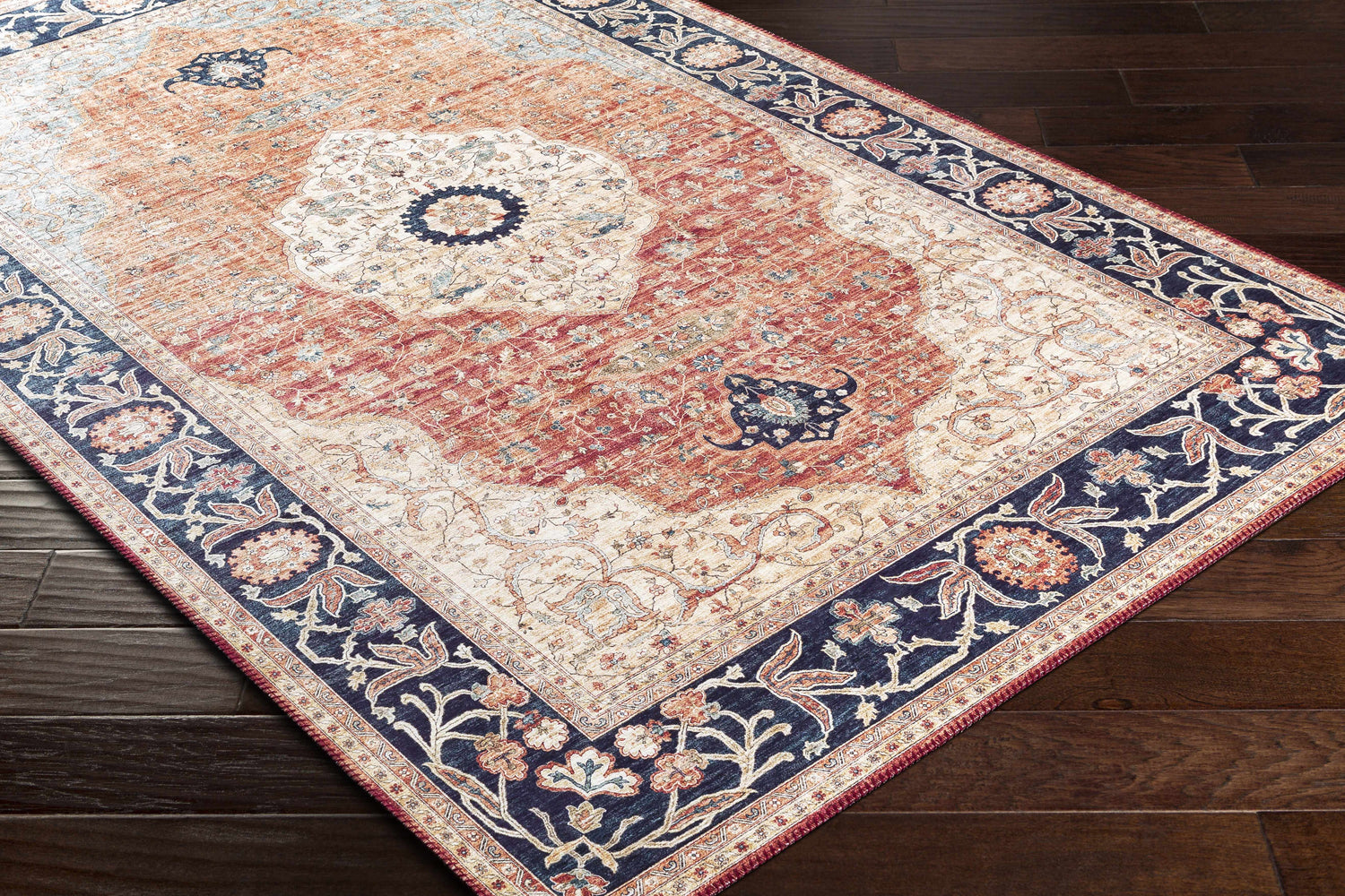 Close-up of the Camperdown Area Rug, a traditional rug in burnt orange, cream, and dark blue on a dark wood floor. Corner