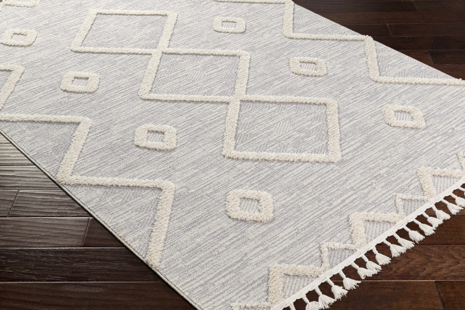 Close-up of the Cauldwell rug. Gray and ivory polyester rug with geometric design, tassels, and high/low pile texture. Corner