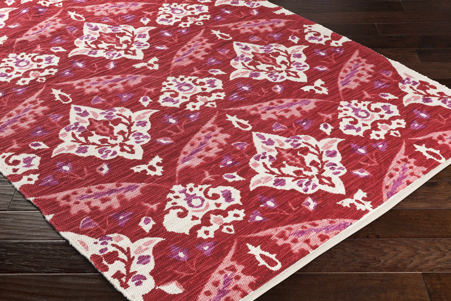 Close-up of the Cook Area Rug, a hand-woven cotton rug with red, pink, purple, and cream floral pattern on dark wood floor. Corner
