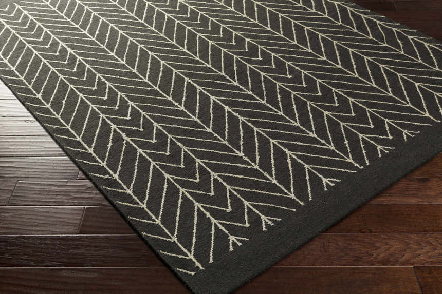 Close-up of the Clymer Wool Rug. Black rug with off-white geometric herringbone pattern on dark wood floors. Corner