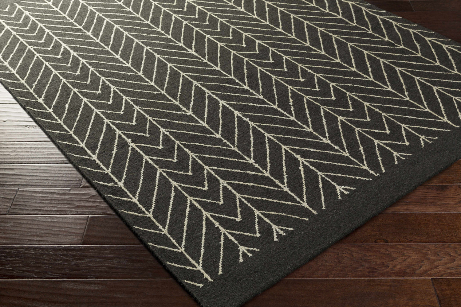 Close-up of the Clymer Wool Rug. Black rug with off-white geometric herringbone pattern on dark wood floors. Corner