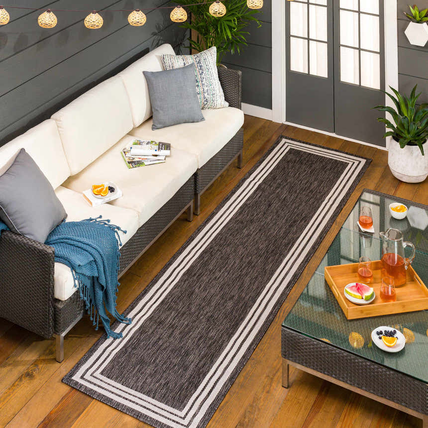 Cozy patio scene with a Coonamble Bordered Antrasit Rug, cream sofa, glass table, and string lights. Perfect outdoor living. Roomscene-Runner