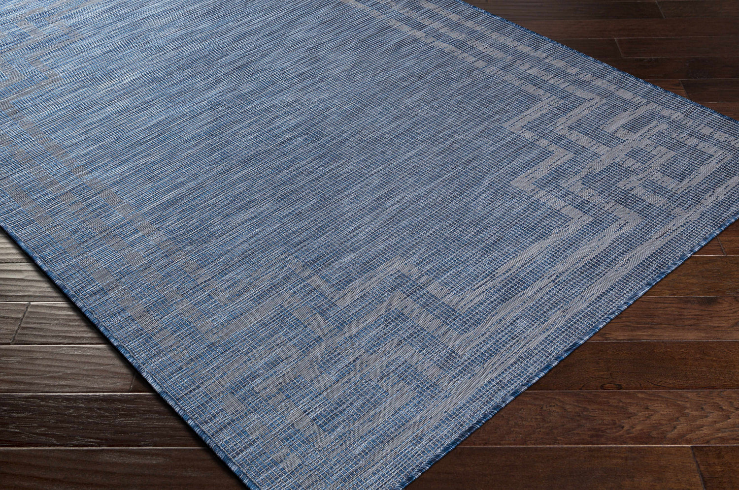 Close-up of the blue Corlette Area Rug showing its woven texture and geometric pattern on a dark wood floor. Corner