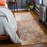 Bedroom scene with a colorful Cranfield Area Rug in brick red, cream, and blue on a hardwood floor. Bed with white and gray blankets. Roomscene-Runner