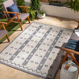 Outdoor patio scene with the Dapitan rug in light gray and denim, surrounded by wooden chairs and potted plants. Roomscene-Rectangle