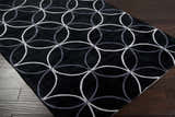 Close-up of the Delano Area Rug. Black rug with light gray and charcoal circle pattern on dark wood floor. Corner