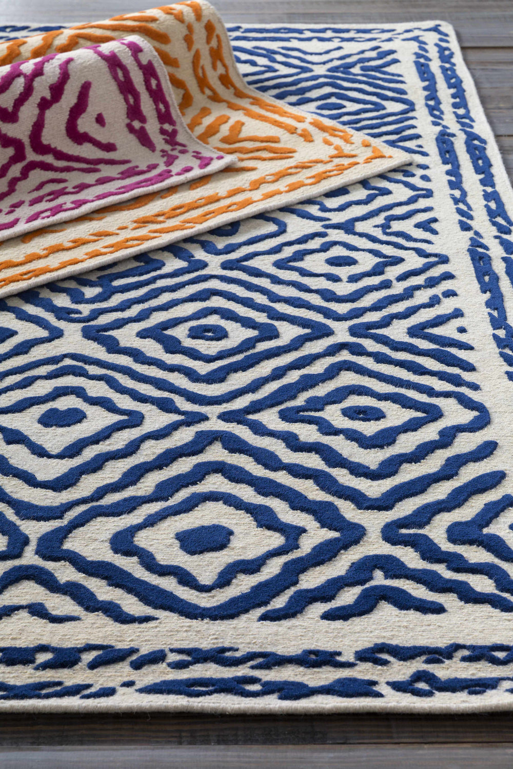 Close-up of Dundalk Area Rug. Hand-knotted wool rug with dark blue geometric pattern on light beige. Other rugs in pink and orange. Styleshot