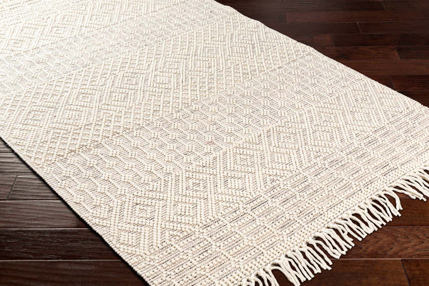 Close-up of Egil Wool-blend Area Rug in off white with a geometric diamond pattern and fringe, on dark wood flooring. Corner