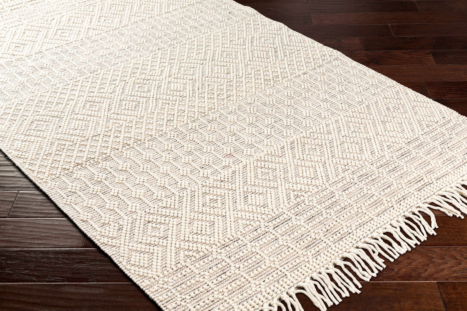Close-up of Egil Wool-blend Area Rug in off white with a geometric diamond pattern and fringe, on dark wood flooring. Corner