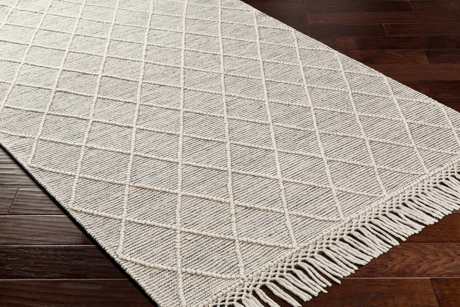 Close-up of Ezria Wool Rug with Tassels showing bone geometric pattern and fringe detail on dark wood floor. Corner