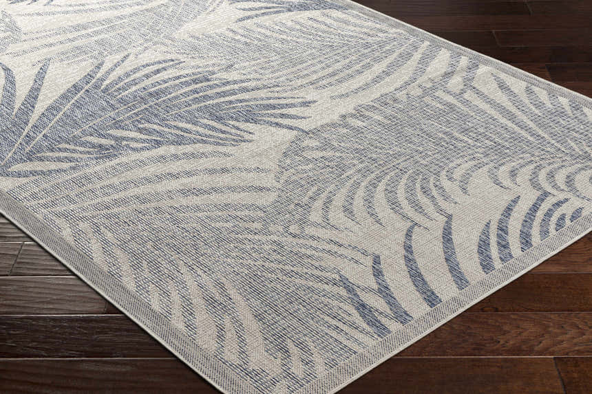 Close-up of Falk Area Rug featuring a cream and navy tropical palm leaf pattern on dark wood floors. Coastal style. Corner