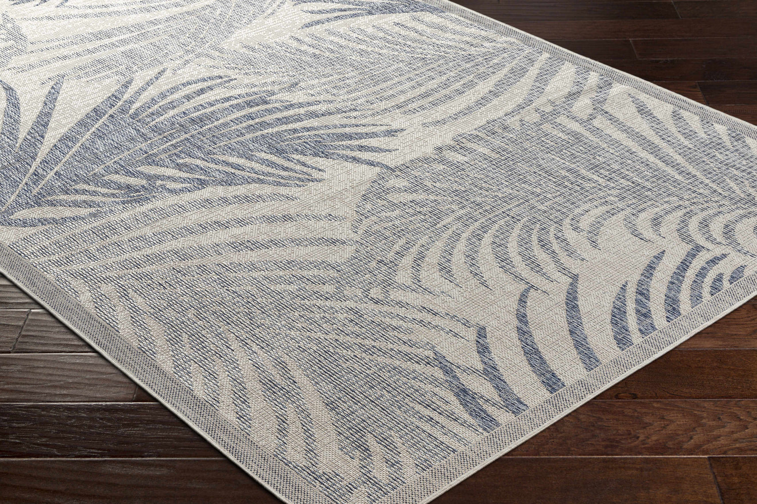 Close-up of Falk Area Rug featuring a cream and navy tropical palm leaf pattern on dark wood floors. Coastal style. Corner