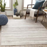 Living room scene with Fombell Abstract Striped Area Rug in charcoal, gray, tan, and white. Modern armchair and blue knit pouf. Roomscene-Rectangle
