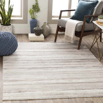Living room scene with Fombell Abstract Striped Area Rug in charcoal, gray, tan, and white. Modern armchair and blue knit pouf. Roomscene-Rectangle