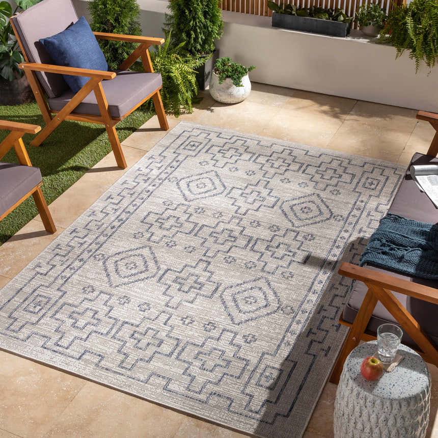 Outdoor patio scene with a Fernway Indoor & Outdoor Rug in blue and taupe, surrounded by wooden chairs and potted plants. Roomscene-Rectangle