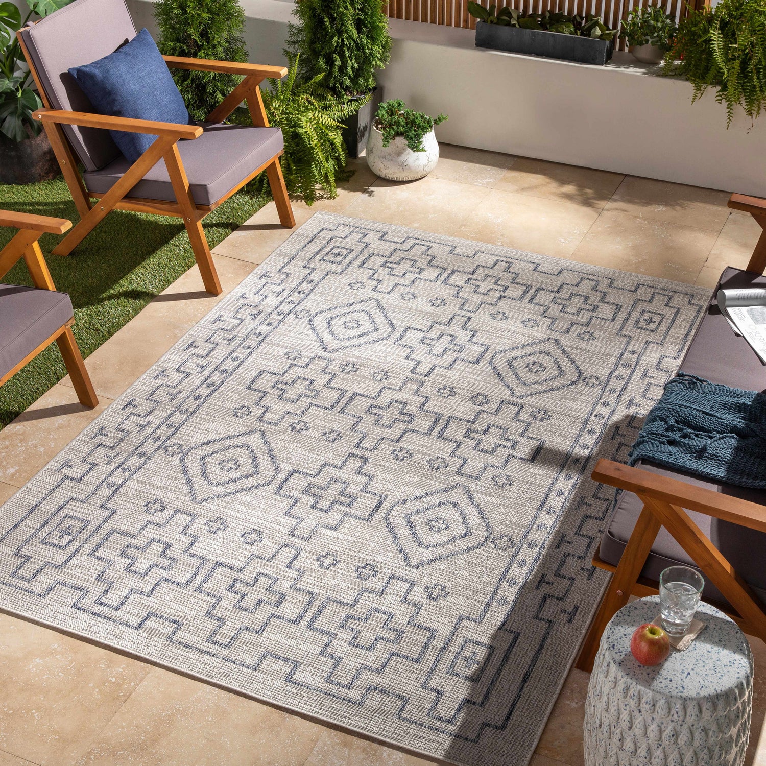Outdoor patio scene with a Fernway Indoor & Outdoor Rug in blue and taupe, surrounded by wooden chairs and potted plants. Roomscene-Rectangle