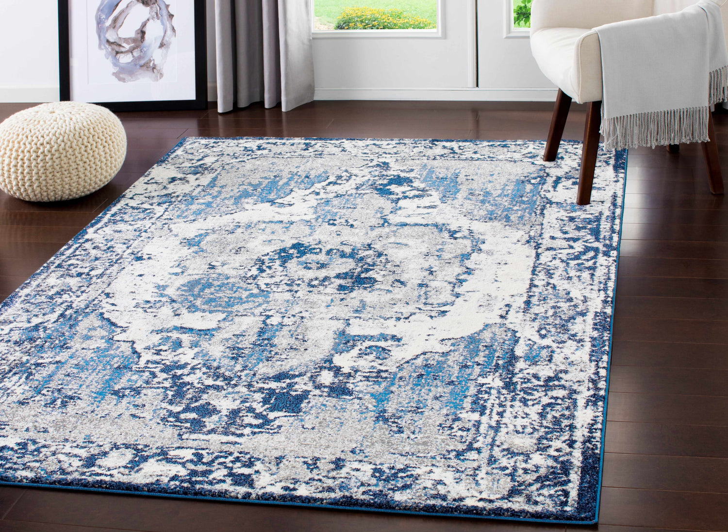 Living room scene with blue and ivory Hartshorne Area Rug, white chair, knitted pouf, and dark wood floors. Roomscene-Rectangle
